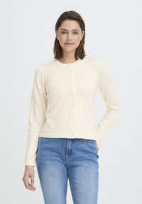 Cardigan à manches longues de couleur crème claire avec un col rond, une fermeture boutonnée sur le devant et une coupe ajustée, associé à un jean bleu. Texture lisse.