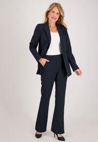 Vrouw met blond haar, gekleed in een marineblauwe blazer en broek, een wit topje, gouden oorbellen en hoge hakken, die staat met één hand in haar zak.