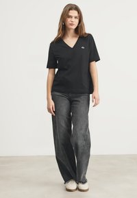 T-shirt nera con scollatura a V e maniche corte, realizzata in tessuto morbido. Abbinata a jeans grigi larghi e sneakers bianche. Presenta un piccolo logo sul petto.