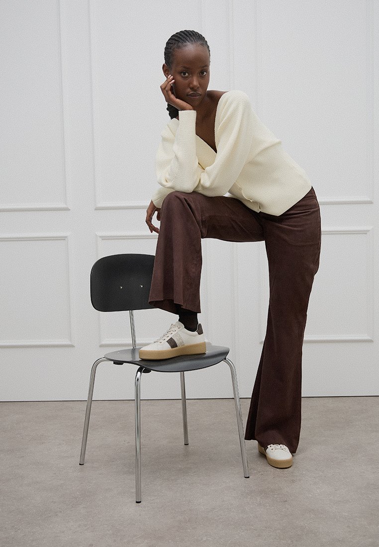 Suéter de punto crema, pantalones amplios marrones, zapatillas blancas con acentos marrones y una silla negra en un entorno interior minimalista.