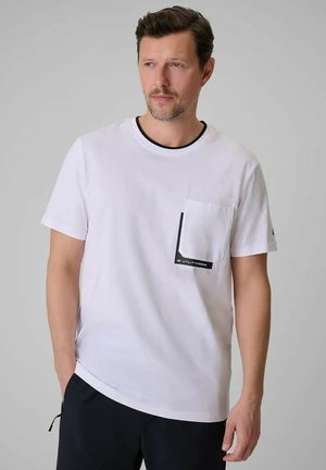 Mężczyzna w białym krótkim rękawem t-shircie z czarnym wykończeniem kieszeni z napisem "UTILITY/WEAR" i czarnych spodniach, stojący na jednolitym szarym tle.