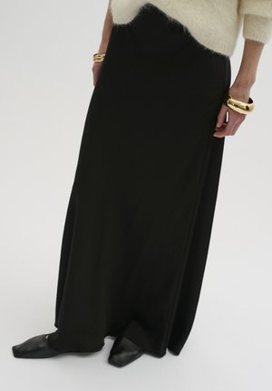 Long black skirt made of smooth fabric, featuring a slight sheen, worn with black slip-on shoes and gold bangle bracelets.
