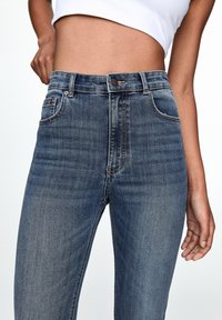 Jeans de mezclilla azul de tiro alto con una textura suave, diseño de cinco bolsillos y costuras en contraste. Cuenta con un cierre de un solo botón.