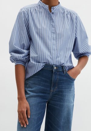 Personne portant un chemisier à rayures verticales bleu et blanc avec des manches bouffantes et un jean en denim bleu, une main dans la poche, sur un fond uni.