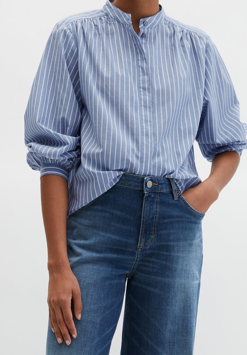 Personne portant un chemisier à rayures verticales bleu et blanc avec des manches bouffantes et un jean en denim bleu, une main dans la poche, sur un fond uni.
