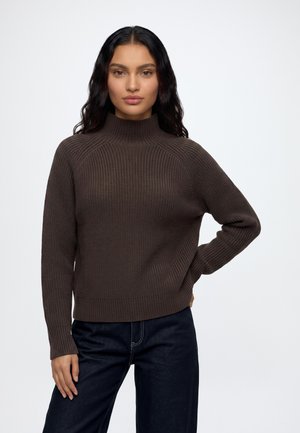 Vrouw met lang donker haar, gekleed in een bruine geribde trui en donkere broek, staand met één hand op haar heup tegen een effen achtergrond.
