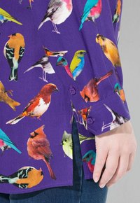 Blusa viola con una vivace stampa di uccelli in vari colori, con polsini abbottonati e spacchi laterali. Il tessuto è leggero e strutturato.