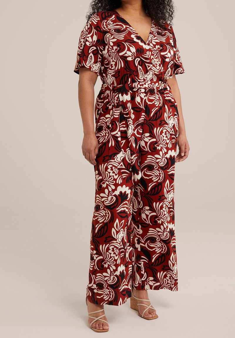 Femme portant une combinaison pantalon à jambes larges avec des motifs floraux rouges, blancs et noirs, et des sandales compensées à brides beiges.