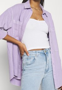 Chemise oversize lavande à boutons avec une poche avant unique, portée sur un top ajusté blanc, associée à un jean en denim bleu clair.