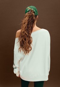 Felpa oversize bianca con design scollato, caratterizzata da maniche lunghe. Abbinata a un cappellino verde e capelli lunghi e mossi.