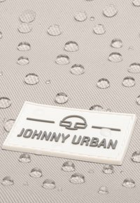 Vízálló bézs anyag, amelyen átlátszó vízcseppek láthatók. Egy fehér címke szürke felirattal olvasható: "JOHNNY URBAN." Texturált felület, minimális dizájnnal.