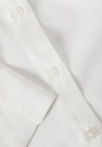 Detail bílé bavlněné košile, která má zakřivený límec, hladkou texturu a tři bílé knoflíky s viditelným prošíváním na léze.
