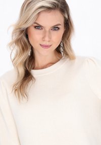 Kobieta z falującymi blond włosami, ubrana w sweter z kremowej dzianiny i srebrne kolczyki w kształcie liści, patrzy pewnie w obiektyw.