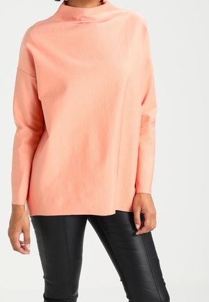 Mujer con una blusa suelta de manga larga color melocotón claro con el cuello alto y pantalones negros ajustados, de pie frente a un fondo liso.