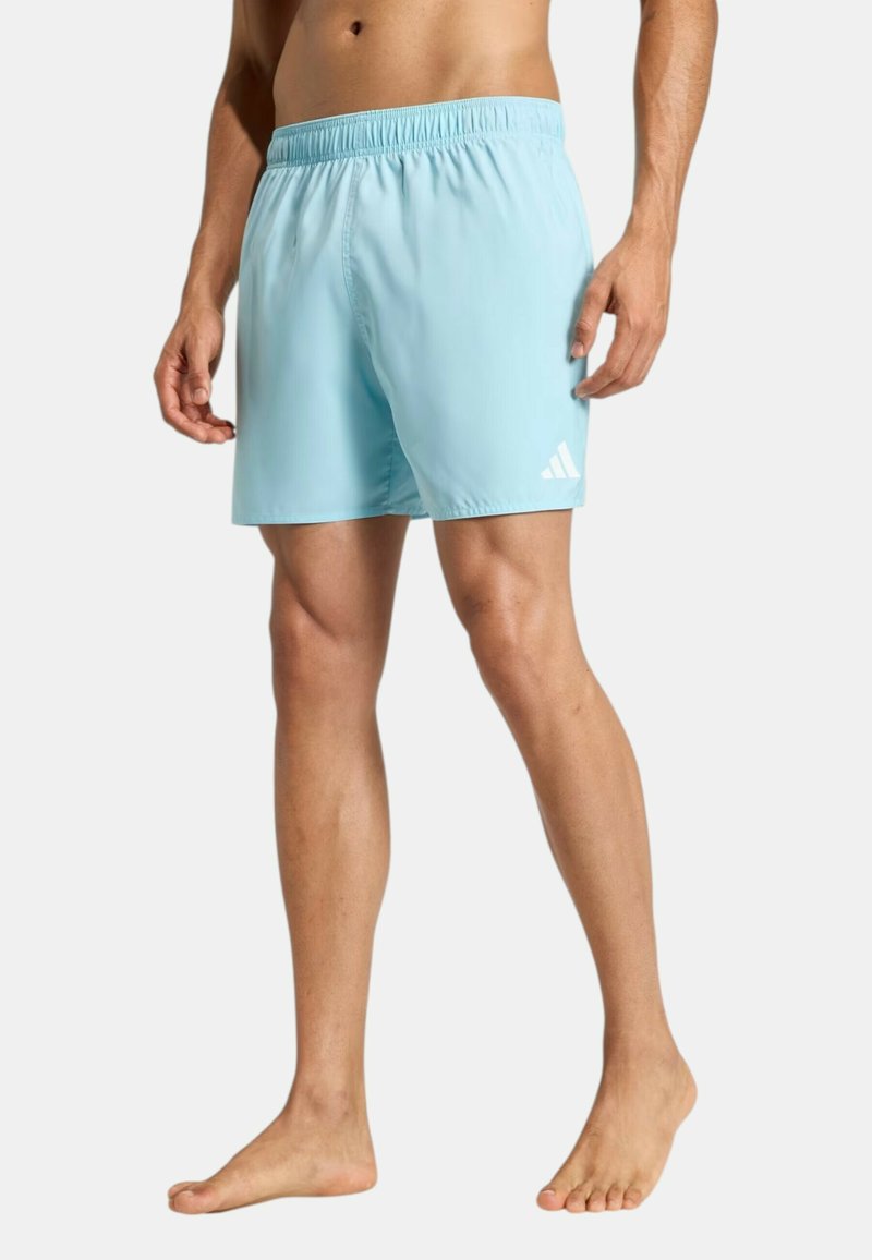 Hombre con pantalones cortos deportivos azul claro con cintura elástica y logo blanco de Adidas en la pierna izquierda, de pie descalzo sobre fondo blanco.