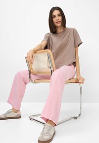 Losse, korte mouwen taupe t-shirt gecombineerd met roze wijde pijpen. Model draagt beige sneakers met roze accenten en zit op een houten stoel.