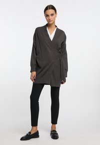 Mörkgrå omslagströja med V-hals, långa ärmar och sidoknytdetalj, i kombination med svarta leggings och loafers. Slät textur.