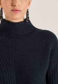 Nahaufnahme einer Person, die einen schwarzen strukturierten Rollkragenpullover und lange schwarze, mit Juwelen besetzte Ohrringe trägt, mit Lippen, die mit einem hellen rosa Lipgloss versehen sind.