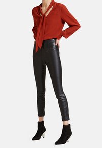 Červená blůza s dlouhými rukávy a detailem vázání na výstřihu, spárovaná s přiléhavými černými koženými kalhotami a černými kotníkovými botami s bílým podpatkem.