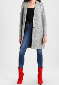Grauer, langer Mantel mit Revers und zwei Knöpfen, kombiniert mit blauen Skinny Jeans und roten, knöchelhohen High Heels. Glatte Textur.