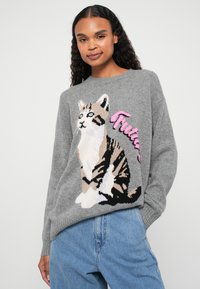 Pull en maille grise avec un motif de chat en beige, noir et blanc, accompagné d'une inscription en rose disant "Future". Coupe décontractée et poignets côtelés.