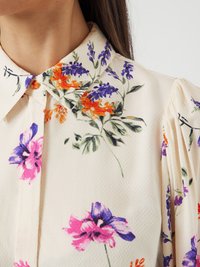 Blomstrete bluse med kremfarget bakgrunn og lilla, rosa og oransje blomster. Krage-design med teksturert stoff og rynkede ermer.