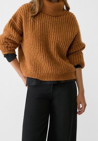 Femme portant un pull à col roulé épais marron sur un chemisier noir à manches longues et un pantalon noir ample, debout devant un fond uni.
