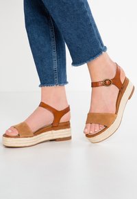 Sandali in pelle marrone e suede con piattaforma, suola intermedia in juta intrecciata, punta aperta, due cinturini e un accento metallico circolare vicino alla caviglia.