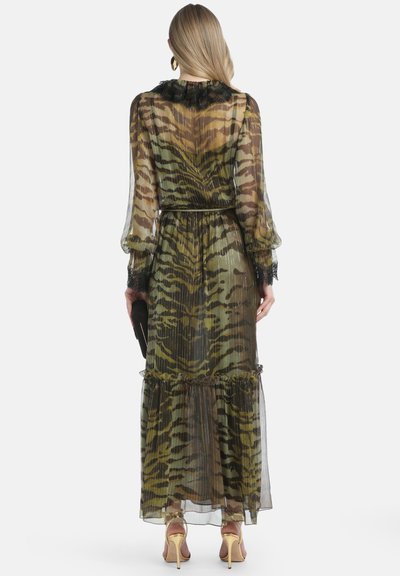 Frau in einem transparenten, langärmligen Kleid mit Tigerdruck, schwarzen Spitzebesätzen und Kragen, hält eine schwarze Clutch und trägt goldene High Heels.