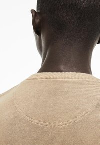 Gros plan de l'arrière de la tête et du cou d'une personne aux cheveux courts, portant un pull en tricot beige.