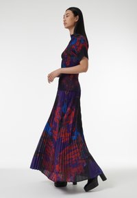 Largo vestido plisado en púrpura oscuro con patrones abstractos en rojo y azul. Parte superior de punto con mangas cortas y silueta ajustada. Tacones de plataforma negros.