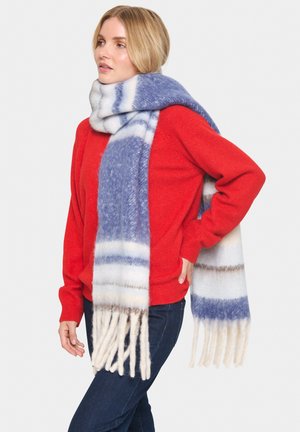 Femme blonde portant un pull rouge, un jean bleu et une longue écharpe rayée bleue, blanche et beige avec des franges aux extrémités.