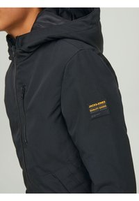 Osoba nosząca czarną kurtkę z kapturem Jack & Jones z zamkniętą kieszenią na piersi i metką marki na górnej części rękawa.