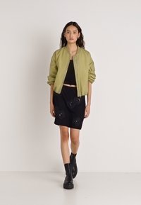 Olijfgroene bomberjack, zwart cropped topje, zwarte rok met bloemrijke borduurwerk en zwarte enkellaarsjes, model staand tegen een effen achtergrond.