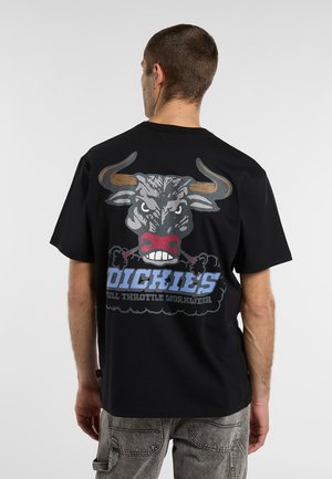 Hombre con camiseta negra con gráfico de toro enfadado y texto "Dickies Full Throttle Workwear" en la espalda, de pie frente a un fondo liso.