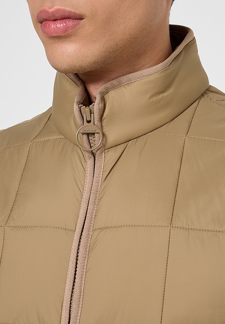 Giacca isolante color sabbia con colletto alto, dotata di chiusura frontale a zip e motivo trapuntato. Texture della superficie liscia con accenti beige.