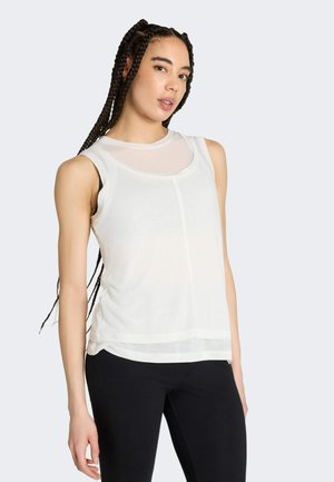 Giovane donna con lunghe trecce che indossa una canotta bianca senza maniche con scollo trasparente e pantaloni neri aderenti, in piedi davanti a uno sfondo chiaro.