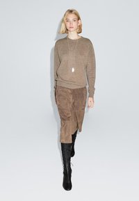 Maglione marrone lavorato a maglia, gonna in suede marrone e stivali neri alti fino al ginocchio. Il modello indossa una collana lunga, toni neutri su uno sfondo grigio chiaro.