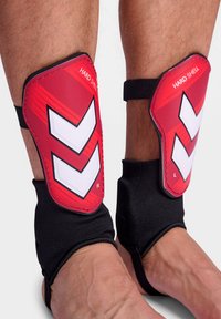 Rode harde scheenbeschermers met witte chevron-accenten. Zwarte elastische mouwen houden ze op hun plaats rond de onderbenen voor bescherming tijdens de sport.
