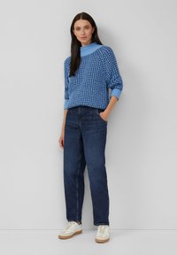 Vrouw met lang, donker haar draagt een blauwe, gebloemde trui, donkere spijkerbroek en witte sneakers, staand tegen een lichtgrijze achtergrond.