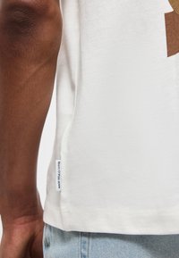 Κοντινό πλάνο του βραχίονα ενός ατόμου που φοράει λευκό μπλουζάκι με μια μικρή ετικέτα «Marc O'Polo Denim» κοντά στο τελείωμα και ανοιχτόχρωμο μπλε τζιν.