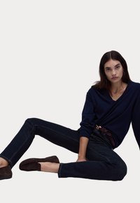 Pull en V marine, jeans en denim foncé et chaussons mocassins marron. Tissu doux, coupe décontractée et détails de surfilage subtils sur les jeans.