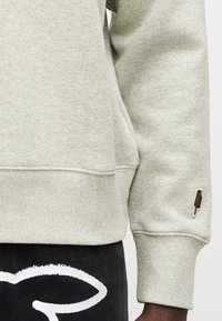 Ανοιχτό γκρι φούτερ με ριμπ στη βάση και στα μανίκια, με ένα μικρό πατς παγωτού στο μανίκι. Φορεμένο με μαύρο παντελόνι που έχει λευκό γραφικό σχέδιο.