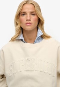 Sweat-shirt beige avec le texte "SUPERDRY" brodé, en tissu doux, col rond, porté par-dessus une chemise à col rayée bleue.