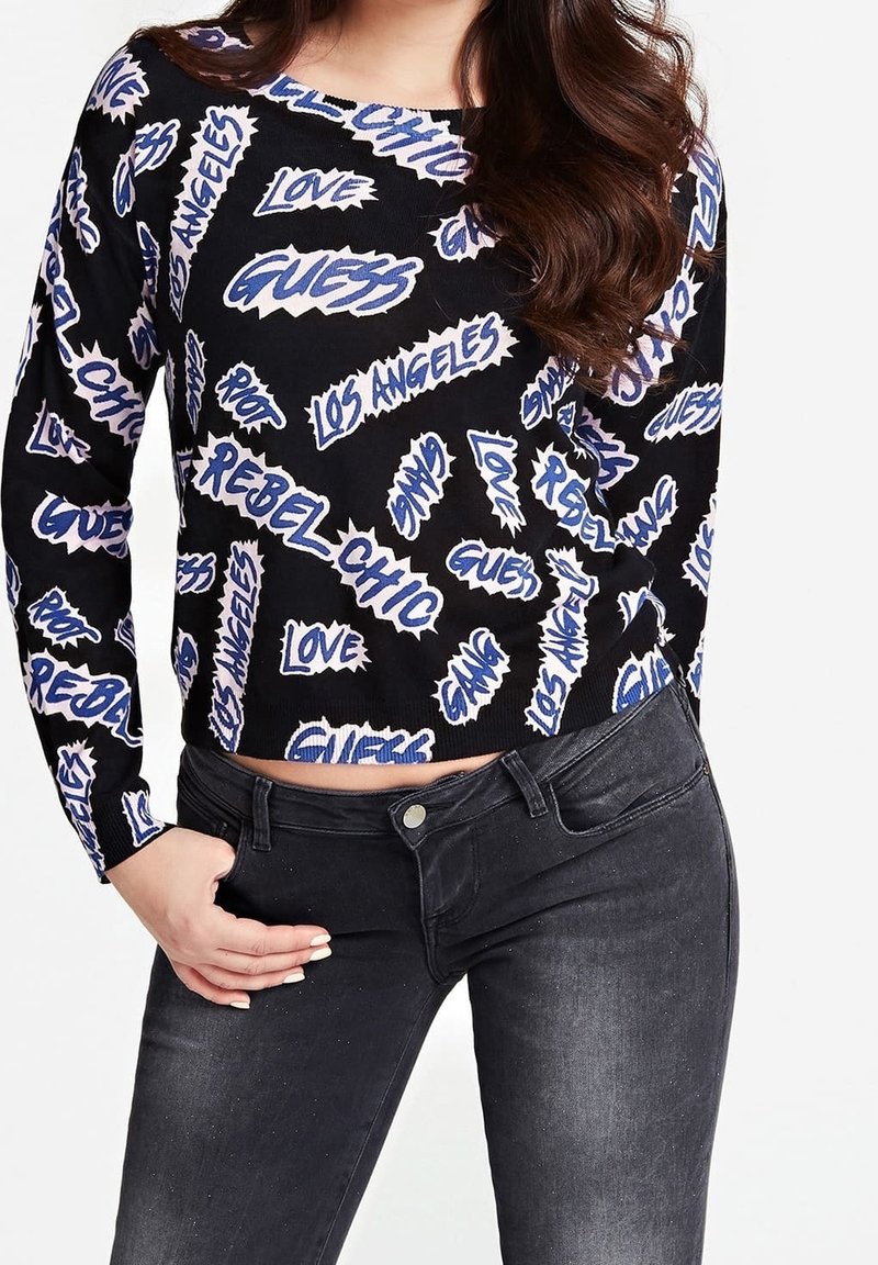 Pull en maille noire arborant un texte graphique vibrant en bleu et blanc, avec une coupe courte. Porté avec un jean foncé, mettant en valeur le détail de la taille.