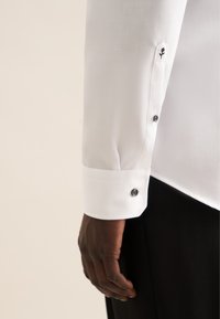 Witte dress shirt met een gladde textuur, met een zwart knoopje op de manchette en een geborduurd bloemenpatroon nabij de knoopsgaten.
