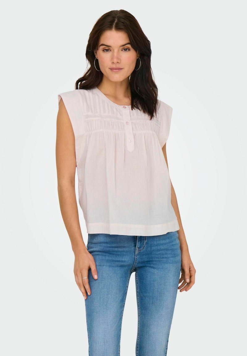 Jasnoróżowa bluzka z krótkim rękawem, posiadająca zapięcie na guziki i marszczenia przy karczku. Połączona z niebieskimi dżinsami, zaprezentowana w studio.