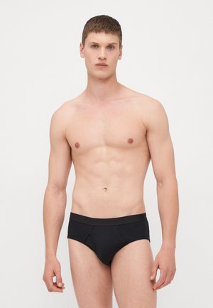 Schwarze Baumwollslips mit breitem elastischem Bund, nahtlosen Seiten und figurschmeichelndem Schnitt, präsentiert auf einem männlichen Model, das vor einem schlichten Hintergrund steht.