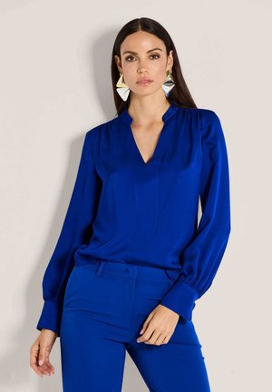 Blaue Bluse mit V-Ausschnitt und langen, voluminösen Ärmeln. Glattes Gewebe mit leichtem Glanz, kombiniert mit passenden blauen Hosen. Silberne geometrische Ohrringe.
