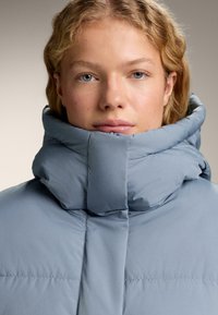 Blondýn s vysokým límcem, v světle modré prošívané bundě na zip, čelí dopředu s neutrálním výrazem na pozadí bez potisku.